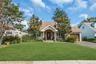 Pitched roof with asphalt shingles, stone facade entrance, attached garage with wood-finish door, paved driveway, and established landscaping