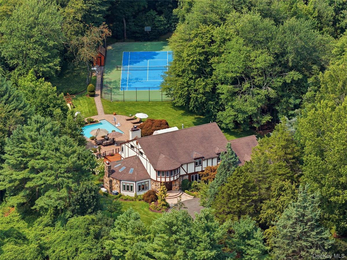 Tudor-style residence set amidst lush trees, featuring mahogany decking, stone patio, tennis court, and swimming pool.