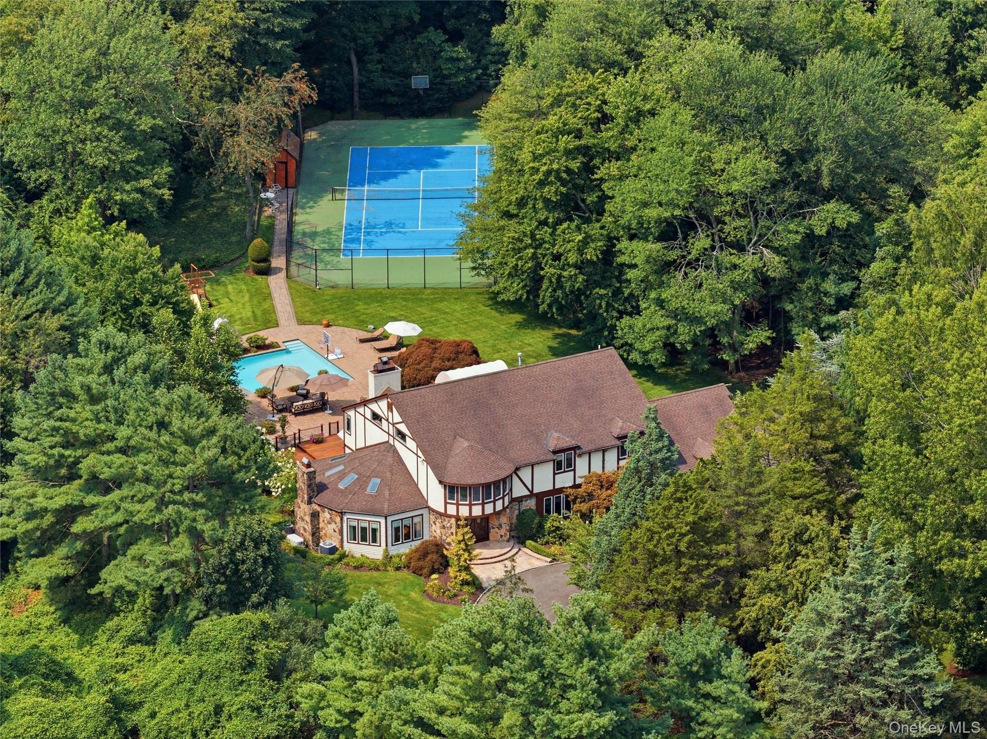 Tudor-style residence set amidst lush trees, featuring mahogany decking, stone patio, tennis court, and swimming pool.