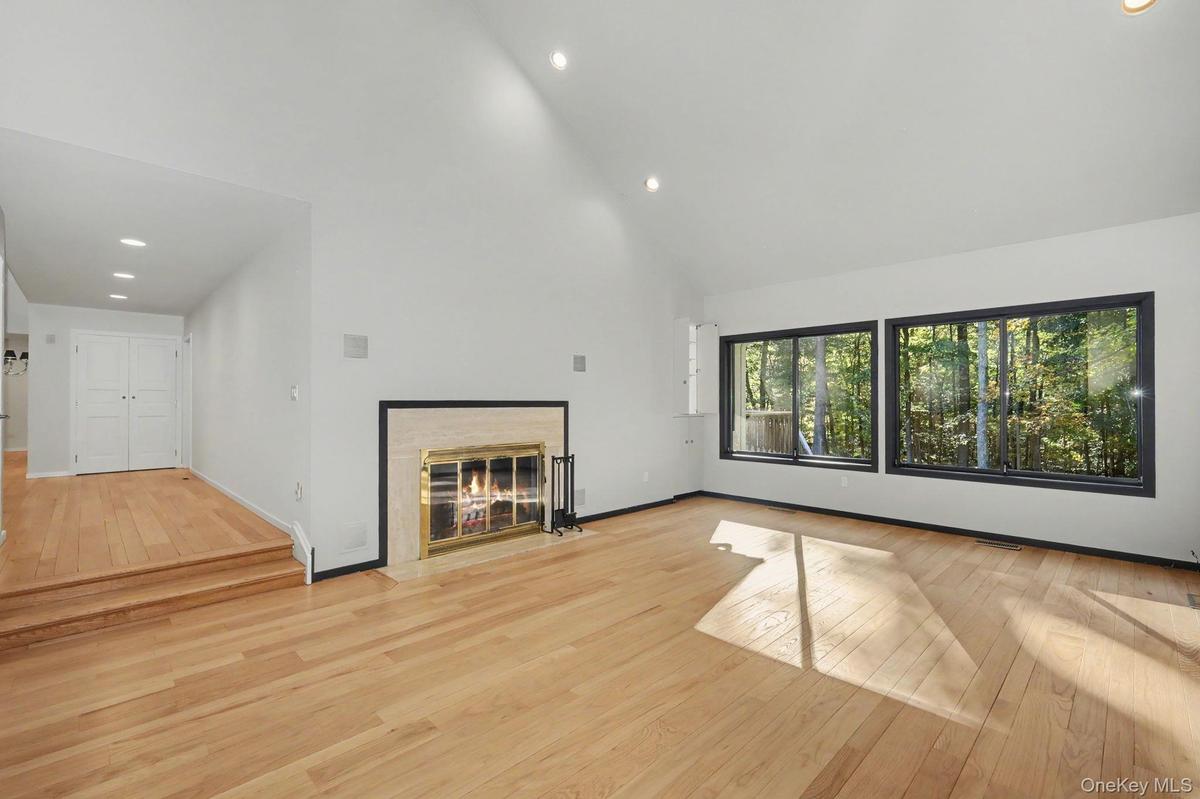 Generous living room with cathedral ceilings, fireplace, and expansive windows overlooking the wooded grounds.