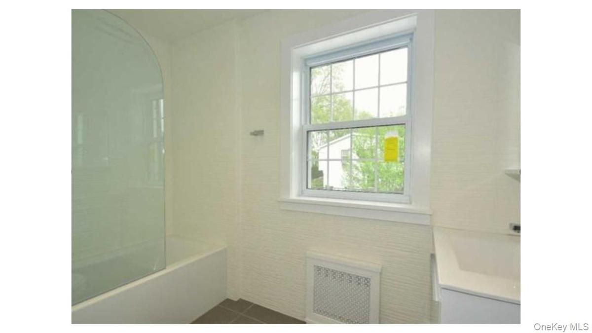 White bathtub with a curved glass shower partition, white window with grid panes, textured wall covering, and a white vanity with an integrated sink