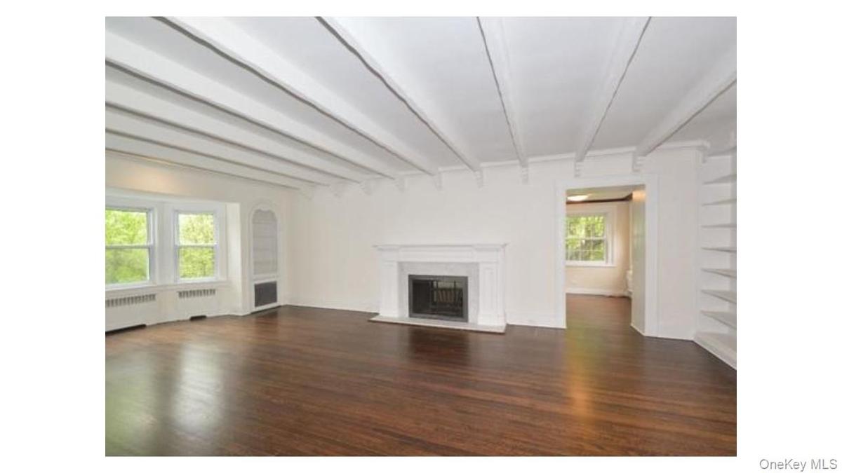 Exposed ceiling beams, rich wood-finish flooring, a classic fireplace with a stone surround, built-in shelving, and a bay window