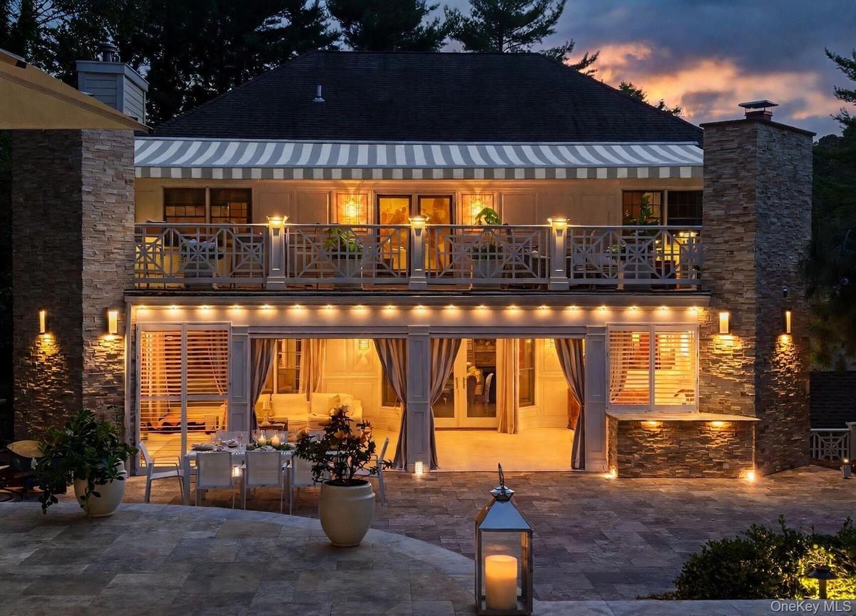 Multi-level exterior with a stone-clad chimney and columns, featuring a second-story balcony with decorative railing, and a retractable striped awning