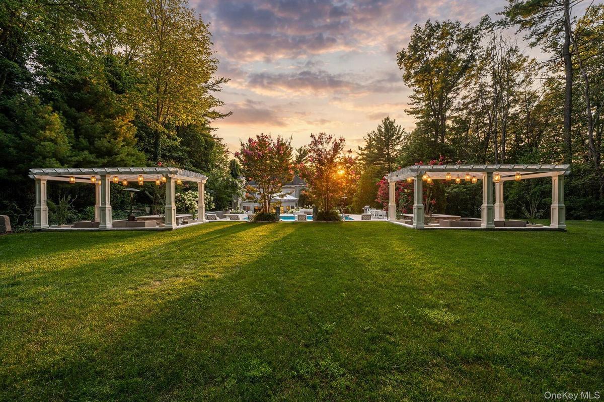 Expansive lawn featuring two white pergolas with integrated lighting, bordered by mature trees and verdant landscaping