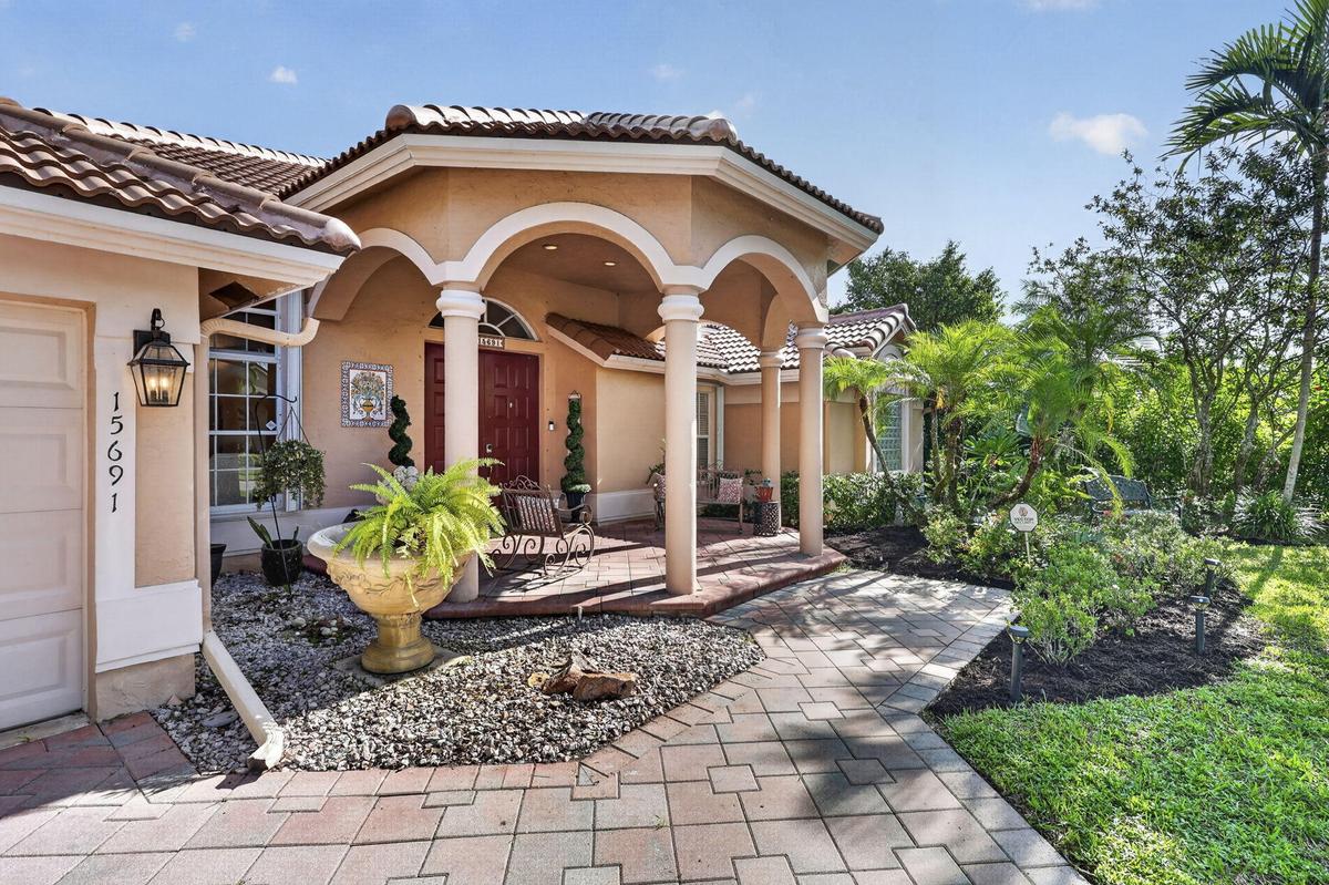 Stucco exterior with a tile roof, featuring an arched portico with columns, a paver walkway, and lush landscaping