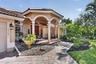 Stucco exterior with a tile roof, featuring an arched portico with columns, a paver walkway, and lush landscaping