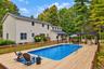 Spacious wood-finish deck with an in-ground swimming pool, a black pergola, and a privacy fence, surrounded by mature trees