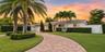 Elegant single-story residence showcasing a curved brick paver driveway, mature palm tree landscaping, and manicured lawn with established hedging