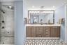 Glass-enclosed shower with marble-style tile, dual vanity with wood-finish cabinetry, stone countertop, and patterned floor tile