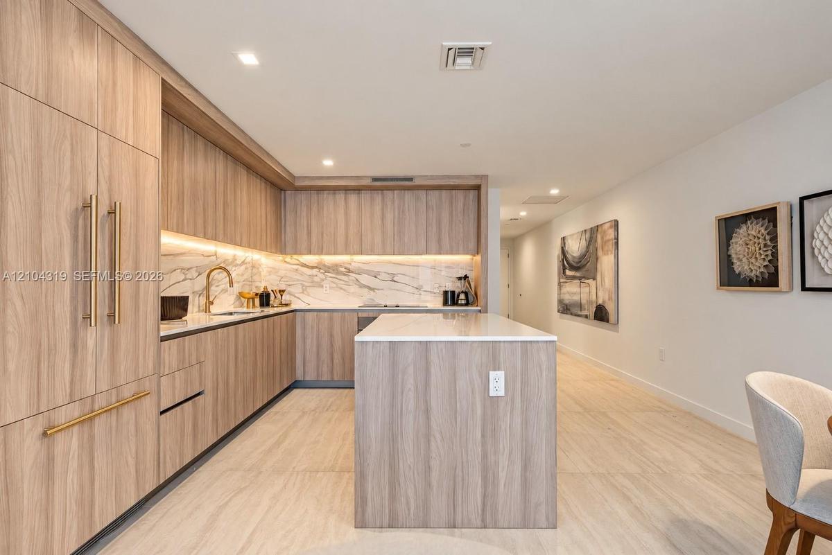Kitchen featuring wood-finish cabinetry, a waterfall-edge island, stone-patterned backsplash, recessed lighting, and metallic hardware