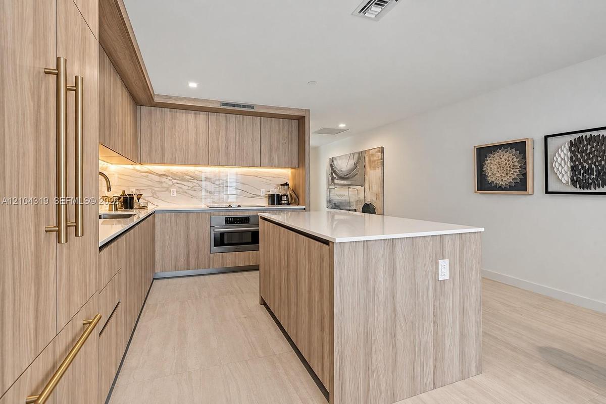 Kitchen featuring wood-finish cabinetry, a large central island with a quartz countertop, integrated under-cabinet lighting, and a stone-style backspl