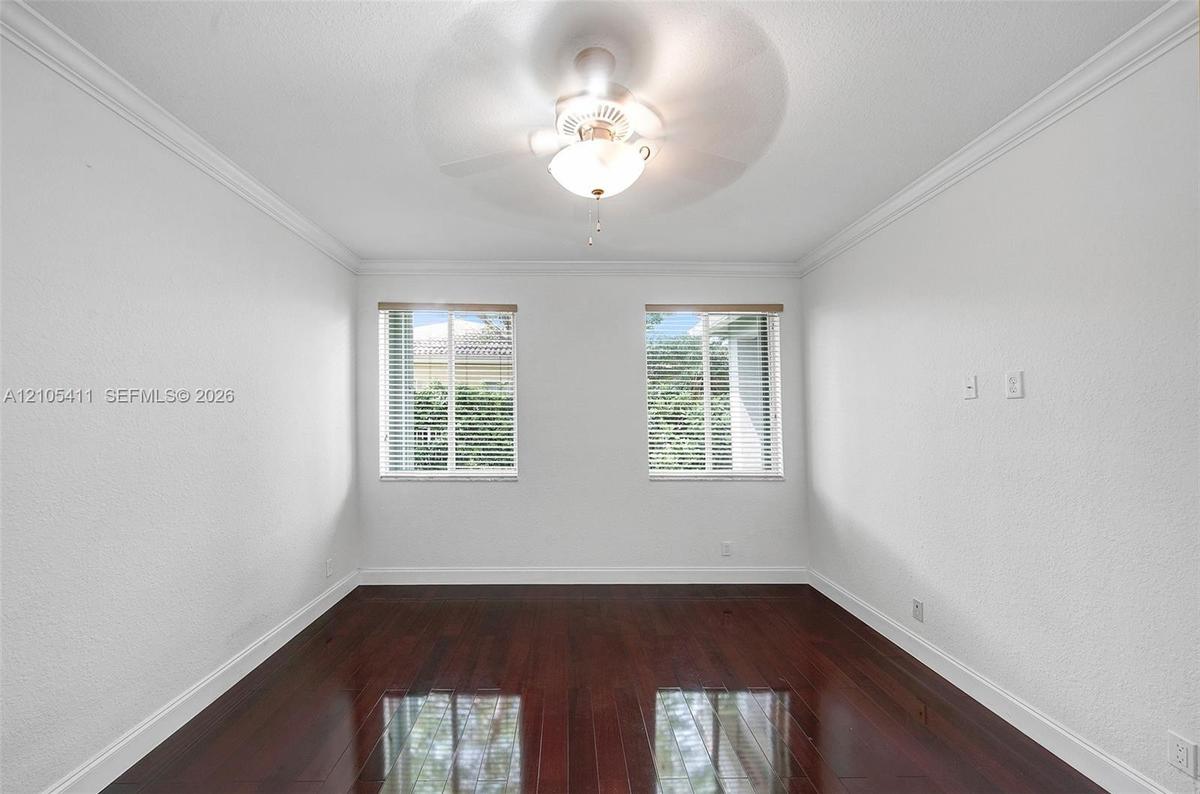 Rich wood-finish flooring, two large windows with blinds, ceiling fan, crown molding, and white textured walls