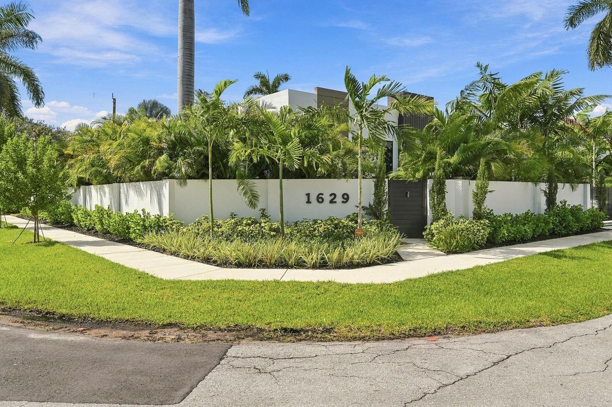 Modern architectural entry featuring a striking white perimeter wall with integrated address signage, lush tropical landscaping including mature palms