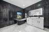 Black-tiled walls with gold veining, a freestanding black soaking tub, a dual vanity with white cabinetry, a light-framed mirror, and large format whi