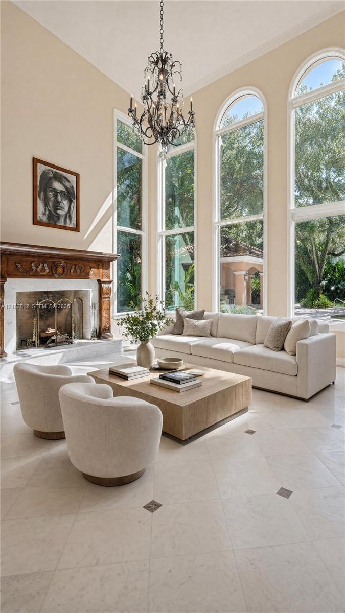 High ceilings featuring an ornate chandelier, a fireplace with a carved wood surround, arched windows, and light-toned tile flooring with dark accent 