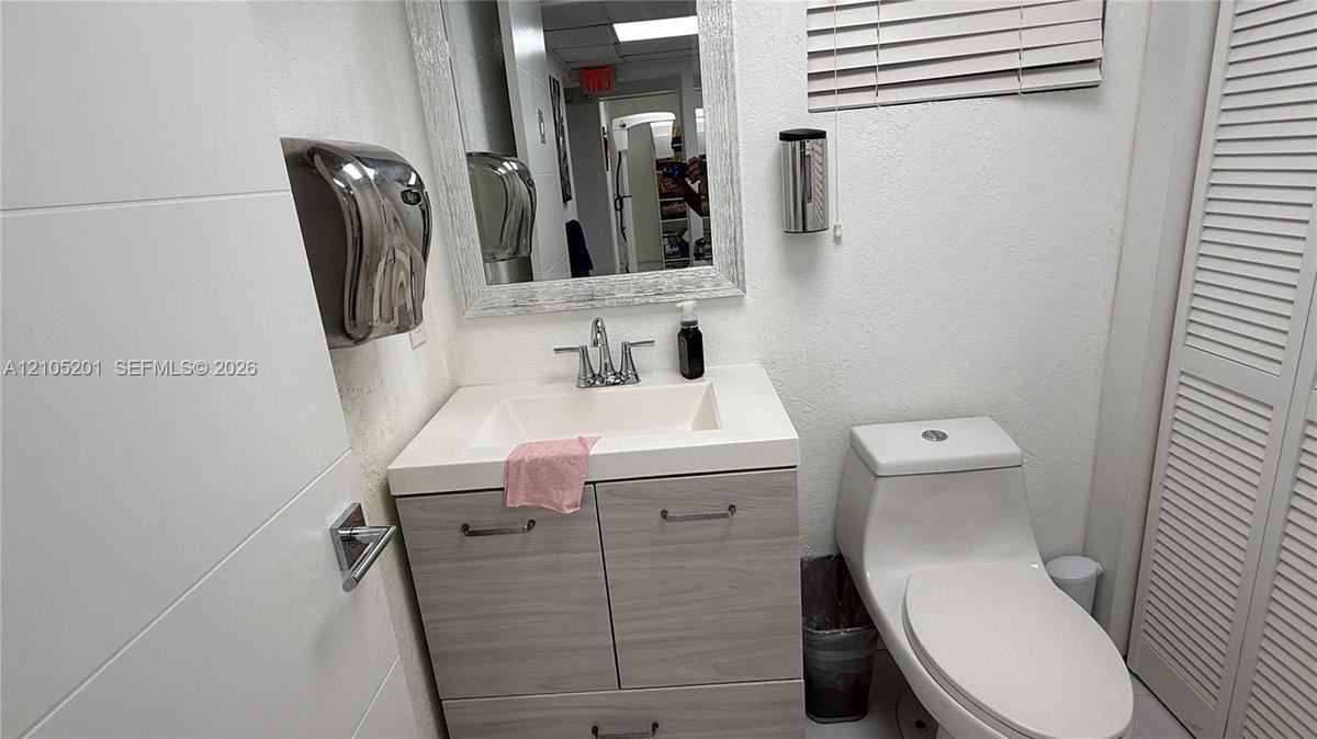 White vanity with wood-finish cabinetry and chrome fixtures, wall-mounted mirror, white wall-mounted toilet, and white louvered bifold doors