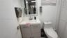 White vanity with wood-finish cabinetry and chrome fixtures, wall-mounted mirror, white wall-mounted toilet, and white louvered bifold doors