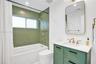 Bathtub with green vertical tile surround, gold-finish shower and bath fixtures, white vanity with green cabinetry, white countertop, and gold-finish 