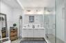 Dual sink vanity with white cabinetry, glass-enclosed shower, light wood-finish flooring, and contemporary wall-mounted light fixtures