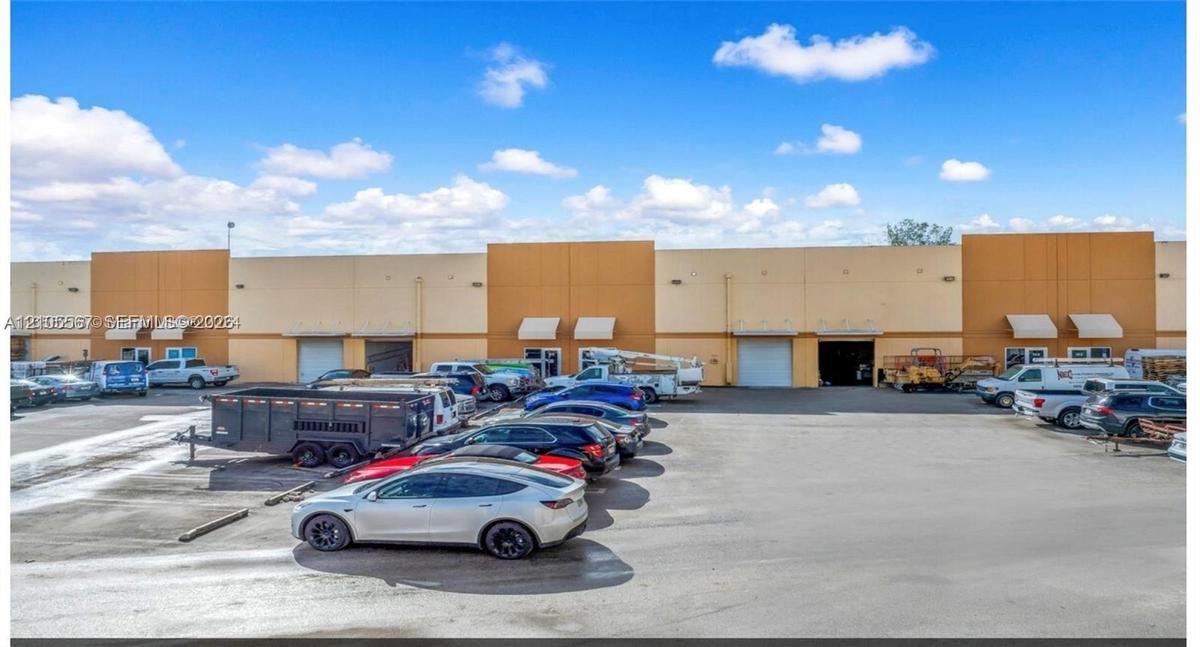 Large paved parking area, commercial building with multiple bay doors, light-colored exterior facade, and exterior awnings