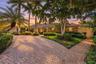 Expansive paver driveway with split path, lush landscaping, and mature palm trees framing a stone-clad exterior