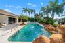 Rectangular swimming pool with rock waterfall feature, surrounding paver patio, and mature tropical landscaping