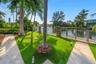 Waterfront property featuring a metal fence with stone pillars, a stone pathway, artificial turf, and mature palm trees