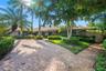 Stone-clad exterior with a tiled roof, paver driveway, and lush landscaping, featuring mature palm trees and established hedging