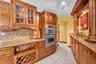 Custom wood cabinetry with glass-front accents, stainless steel double wall ovens, a built-in wine rack, and a stone-finish backsplash with countertop