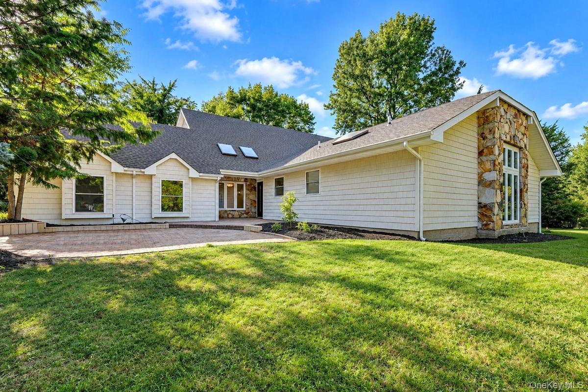 Exterior featuring light-toned shingle siding, natural stone accents, a brick paver patio, roof skylights, and a manicured lawn