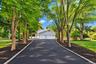 Asphalt driveway bordered by stone pavers, flanked by mature trees and manicured lawns, leading to a two-car garage
