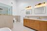 Double vanity with warm wood-tone cabinetry, brass-finish hardware, quartz countertops, dual rectangular undermount sinks, and wall-mounted light fixt