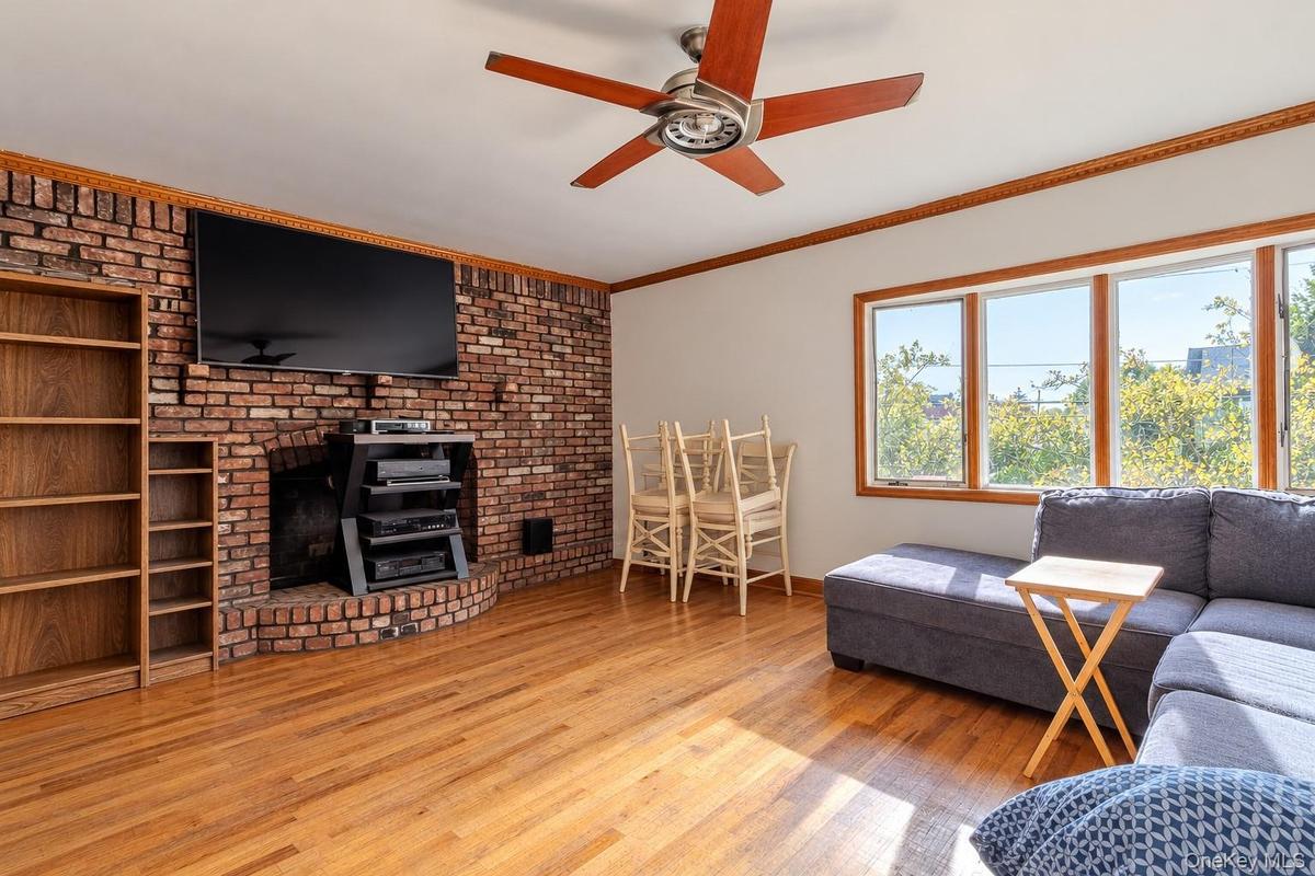 Exposed brick fireplace, wood-finish flooring, ceiling fan, built-in shelving, and large windows with wood-trim casing