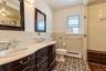 Bathroom featuring a dual-sink vanity with light-toned countertop, ornate dark-finish cabinetry, decorative tile flooring, and a tiled shower surround