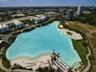 Large-scale recreational lagoon featuring sandy beach shorelines, palm landscaping, paved perimeter walking paths, and expansive grassy grounds