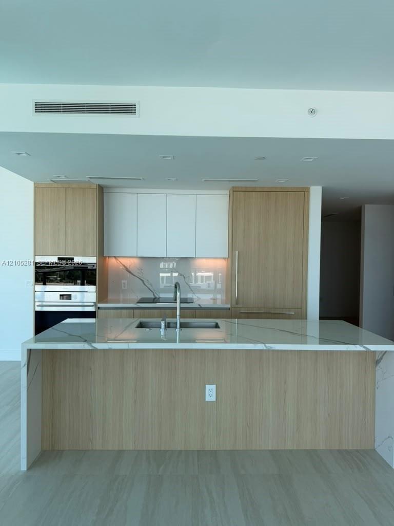 Kitchen featuring wood-finish cabinetry, waterfall island with stone countertop, integrated refrigerator, built-in double oven, and stone-finish backs