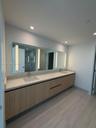 Floating vanity featuring light wood-finish cabinetry, a full-width mirror with integrated LED lighting, and a dual-sink stone-composite countertop