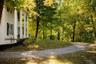 Two-story clapboard exterior featuring a covered front porch, structural columns, shuttered windows, and a gravel driveway set amidst mature trees