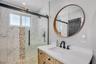 Bathroom featuring a wooden vanity with a white countertop, circular wood-framed mirror, and a walk-in shower with a sliding glass door, patterned sto