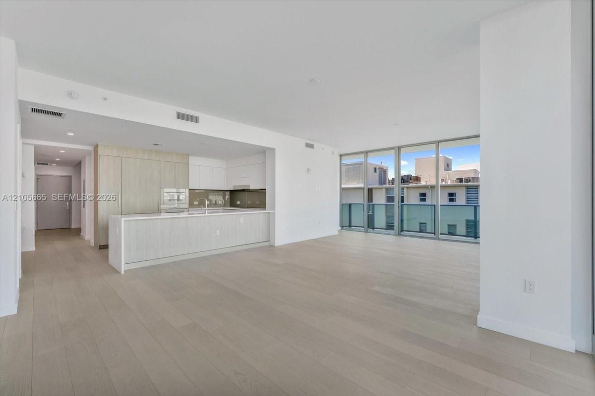 Open-concept interior featuring light-toned wood-finish flooring, floor-to-ceiling glass paneling, and a kitchen equipped with a large island, flat-pa