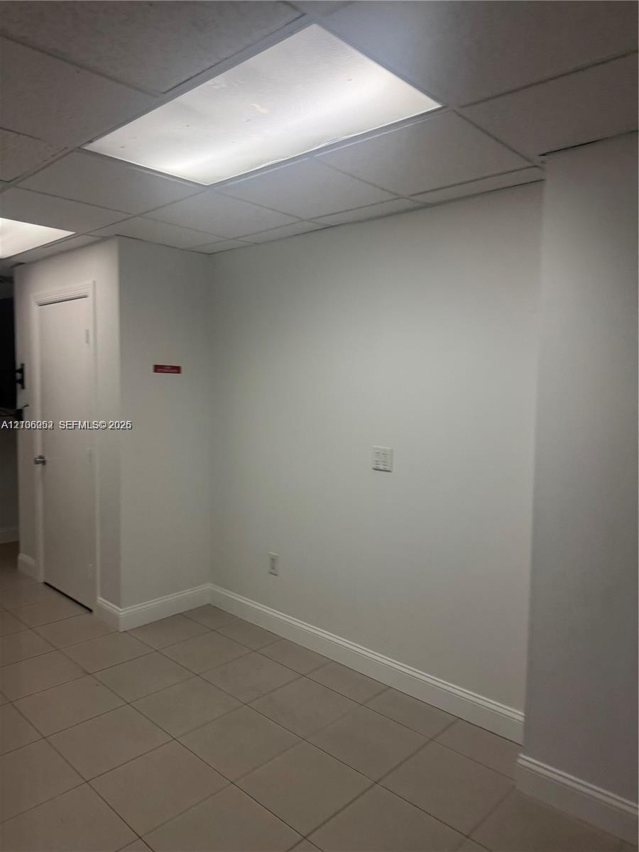 Neutral-toned interior space featuring grid-pattern drop ceiling, recessed lighting panels, ceramic tile flooring, and solid-core interior door