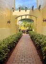 Brick paver walkway lined with manicured hedges, featuring a stucco archway, decorative wood-beam accents, and wall-mounted exterior sconces