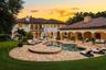Stucco exterior with multi-level tile roofing, expansive stone-paved pool deck, freeform swimming pool with raised spillover spa, and covered loggia f