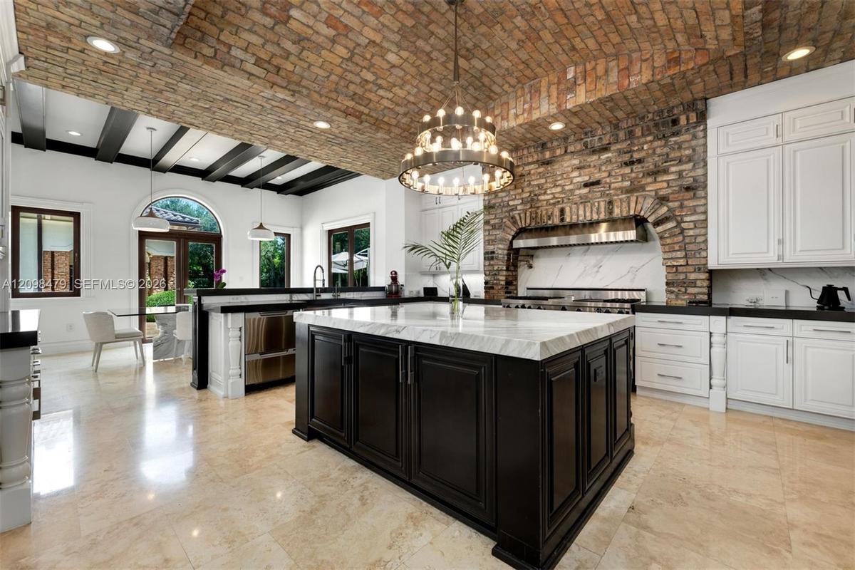 Exposed brick vaulted ceiling with recessed lighting, large central kitchen island with light-colored countertop, white cabinetry, dark stone countert