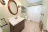 Dark-finish wooden vanity with a white countertop and integrated sink, neutral tile flooring, oval wall mirror, and dual wall-mounted light fixtures