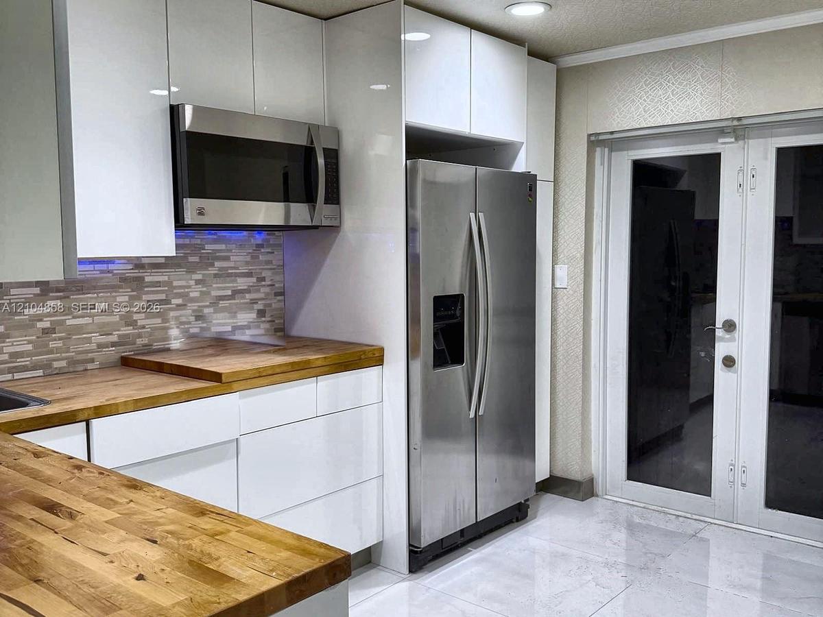 Kitchen featuring high-gloss white cabinetry, wood-finish countertops, stainless steel appliances, multi-tonal tile backsplash, and French doors