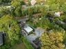 Aerial perspective of the property featuring a gravel driveway, lush green landscaping, mature trees, and a dark gray exterior home
