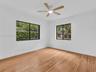 Large front/east bedroom with sunny windows. Featuring wood-finish flooring, two windows with black frames, and a multi-blade ceiling fan with integra