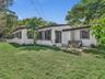 Exterior featuring white stucco siding, dark-trimmed windows, a concrete patio, and a fenced grass lawn with mature trees and palm foliage. House for 
