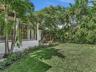 Expansive fenced backyard featuring a manicured lawn, established palm trees, mature greenery, and a concrete patio area. House for rent in Coral Gabl